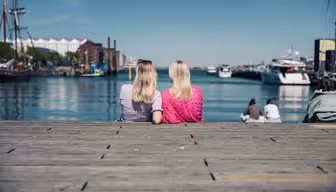 Two people with blonde hair are sitting on a wooden dock by a calm waterfront. They are facing away, one in a purple shirt and the other in pink. Boats and ships are moored in the water, and a couple of people are sitting nearby enjoying the view.