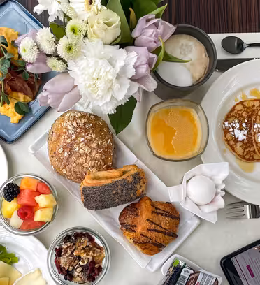 A breakfast spread featuring scrambled eggs with bacon, assorted pastries, pancakes with powdered sugar, a fruit bowl, granola, orange juice, a cappuccino, and a phone. A floral centerpiece decorates the table.