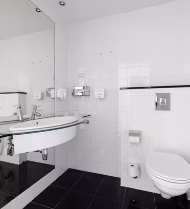 Modern bathroom with a wall-mounted toilet, glass shower enclosure, large mirror, and a floating sink on a black tiled floor. White tiled walls have various fixtures like soap dispensers and a towel holder.