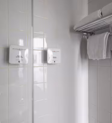 A shower area with two white soap dispensers mounted on tiled walls. A chrome towel rack holds several neatly folded white towels, with a couple hanging on a bar. The tiles are light gray with a grid pattern.