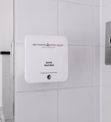 Wall-mounted hand wash dispenser in a tiled bathroom, labeled Gentle Hand Wash. It features the Arp-Hansen Hotel Group logo. A tissue box is partially visible on the right, and two glass containers are on the left.