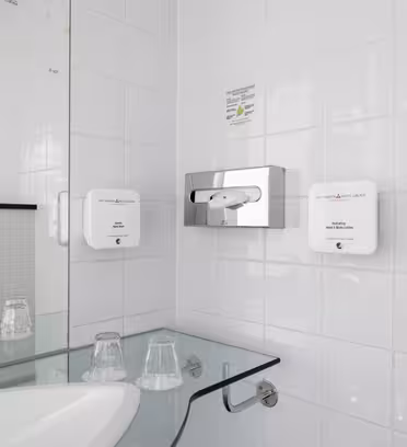 A bathroom with a white tiled wall featuring a tissue holder, two soap dispensers, and a glass shelf holding two plastic cups. A white sink is partially visible in the foreground.