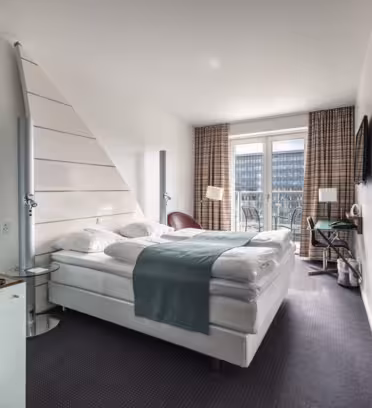 Modern hotel room with a large bed featuring white linens and a blue throw. The room includes a striped wall cabinet, side tables, a flat-screen TV, balcony access, and a round chair by the window. A beverage tray is on a small glass-topped fridge.