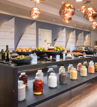 A buffet setup with a variety of food items. The counter features fruits, cereals, and condiments in large jars. Modern pendant lights hang above, and sleek decor with wooden floors and gray walls completes the setting.