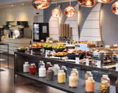 A buffet setup with various foods, including fruits, pastries, and cereals. Glass jars filled with different ingredients line the front. Warm lighting overhead enhances the modern and inviting atmosphere.