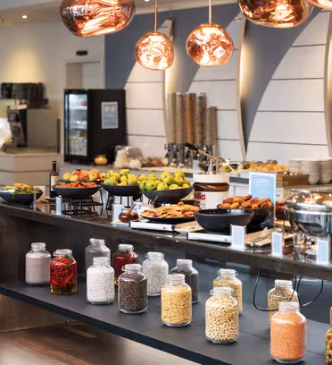 A buffet setup with various foods, including fruits, pastries, and cereals. Glass jars filled with different ingredients line the front. Warm lighting overhead enhances the modern and inviting atmosphere.