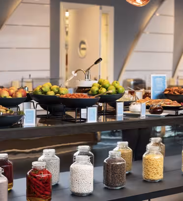 A buffet setup featuring bowls of apples, pears, and various dishes on a table. Below, there are jars filled with red peppers, beans, pasta, and seeds. The background has a modern, sleek design with warm lighting.