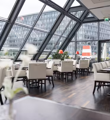 Modern restaurant interior with white chairs and tables on a dark wooden floor. Large slanted windows offer a view of an adjacent office building. Red lamp shades and white flowers add pops of color. The space is empty and well-lit.