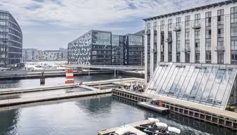 Modern waterfront scene with sleek buildings featuring large windows, a bridge, and a striped lighthouse. Small boats are docked in the foreground, and a partly cloudy sky completes the background.