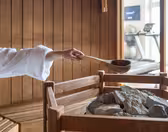 A person wearing a white robe pours water with a wooden ladle over hot stones in a wooden sauna. Treadmills are visible in the background, indicating a spa or fitness facility.