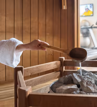 A person wearing a white robe pours water from a wooden ladle onto hot stones in a sauna. Wooden benches line the walls. In the background, there are treadmills and a TV on the wall.