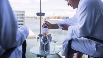 A person in a white robe is sitting on a balcony and opening a bottle of wine or champagne. The bottle is in an ice bucket on a small glass table with two empty wine glasses. A scenic waterfront view is visible in the background.