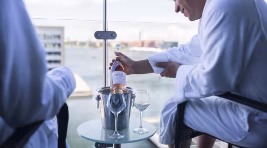 A person in a white robe is sitting on a balcony and opening a bottle of wine or champagne. The bottle is in an ice bucket on a small glass table with two empty wine glasses. A scenic waterfront view is visible in the background.