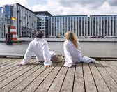 A man and woman sit on a wooden deck by a waterfront, wearing white robes. They face away, towards a modern building with large windows across the water. A wicker basket is beside them under a partly cloudy sky.