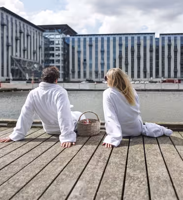 A man and woman sit on a wooden deck by a waterfront, wearing white robes. They face away, towards a modern building with large windows across the water. A wicker basket is beside them under a partly cloudy sky.