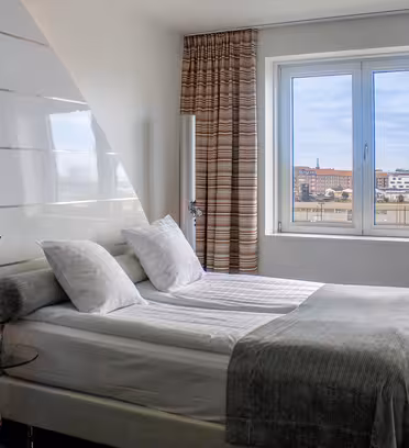 Modern hotel room with a double bed, white linens, and gray blanket. Large window with striped curtains lets in daylight, showing a cityscape outside. Room features minimal decor and a red chair.