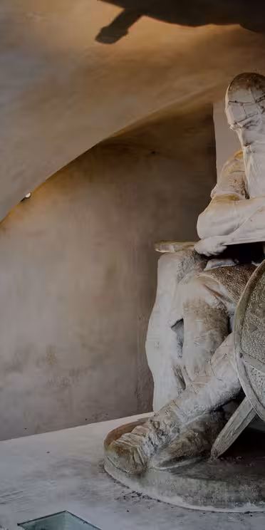 A stone statue of a seated Viking warrior, resting with his arms crossed on his knee. He wears a cape and helmet, and a round shield leans against him. The setting is a dimly lit room with arched ceilings.