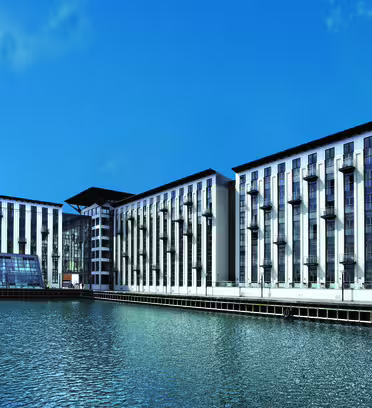Modern waterfront building with a sleek, angular design and large windows, set against a clear blue sky. The structure includes multiple stories and sits beside a calm body of water, reflecting its mirrored surface.