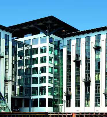 A modern multi-story building with a combination of glass and white facade, featuring numerous windows and small balconies. The structure has a central glass atrium and is set against a clear blue sky.