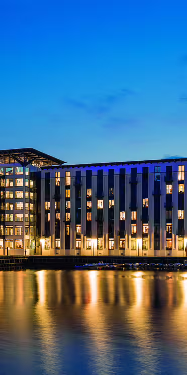 A modern building with large windows and a glass façade is illuminated at night, reflecting on the calm water in front. The sky is twilight blue, enhancing the serene ambiance of the scene.