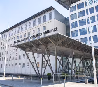 The image shows a modern building labeled Hotel Copenhagen Island. The facade is mostly white with large windows and a steel-framed entrance canopy supported by diagonal beams. The sky is clear.