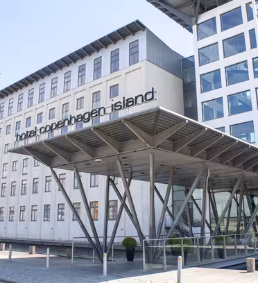 The image shows a modern building labeled Hotel Copenhagen Island. The facade is mostly white with large windows and a steel-framed entrance canopy supported by diagonal beams. The sky is clear.