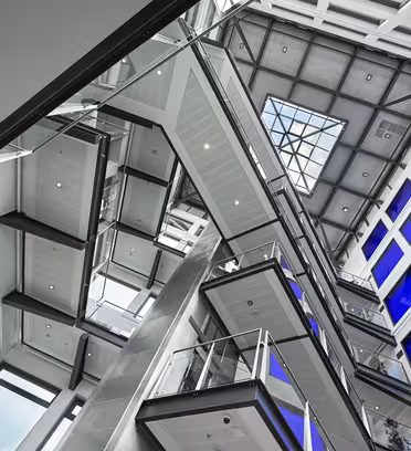 Interior view of a modern buildings atrium, featuring multiple levels of glass balconies and staircases. Large blue-tinted windows line the walls, and a glass ceiling allows natural light to flood in. The design is angular and contemporary.
