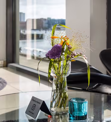 A small bouquet of colorful flowers in a glass vase sits on a glass table in a modern room with large windows. Sunlight illuminates the scene, and two black chairs are visible in the background. A teal candle holder is beside the vase.