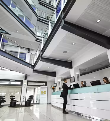 A modern hotel lobby with sleek white and turquoise decor. A person is checking in at the front desk where two staff members assist. Overhead, there are multiple floors with glass railings and contemporary lighting.