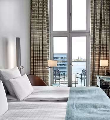 A modern hotel room with two neatly made beds, white and light blue bedding, a large window with a view of water and buildings, two chairs on a small balcony, a wall-mounted TV, and a glass desk with a lamp.