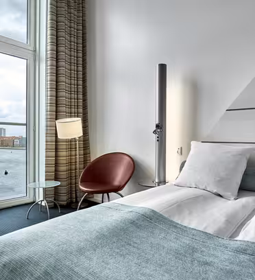 Modern hotel room with a large window showcasing a water view. The space features a neatly made bed with white and light blue bedding, a round red chair, a lamp, and a small glass table next to the chair. The sky is cloudy outside.
