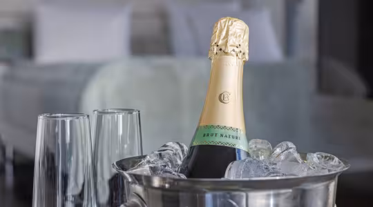 A bottle of Brut Nature champagne chilled in a silver ice bucket, accompanied by two empty champagne flutes. The background shows a blurred view of a neatly made bed with white pillows and a light green bedspread.