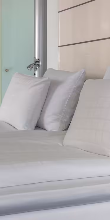 A neatly made bed with white pillows and a white and light blue blanket. In the background, a wooden shelf with glass shelves holds a coffee machine, cups, and a water bottle.