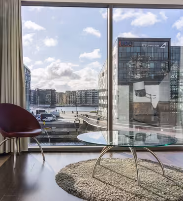 Modern living room with a glass coffee table on a round rug, a maroon chair, and a beige sofa. Floor-to-ceiling window offers a view of urban buildings and a river under a partly cloudy sky.