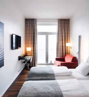 A modern hotel room with two neatly made single beds featuring white linens and gray throws. The room has large windows with curtains, a red armchair, a wall-mounted TV, a desk, and a painting of water ripples on one wall.