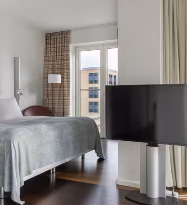 Modern hotel room with a neatly made bed, large flat-screen TV, and floor-to-ceiling window offering a view of a waterfront and nearby buildings. The room features white walls, wooden floors, and soft lighting.