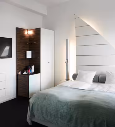 Modern hotel room featuring a large bed with white linens and a teal throw blanket. A flat-screen TV is mounted on the wall. A sleek cabinet with a minibar and modern lighting is in the corner. The décor is minimal with white and wood accents.
