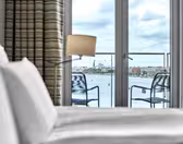 View from a hotel room with an unmade bed in the foreground, looking out through glass doors onto a balcony with two chairs. Beyond the balcony, theres a body of water, industrial buildings, and a cloudy sky in the distance.