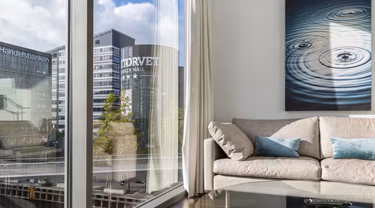 A modern living room with a light gray sofa and blue cushions, adjacent to a large window showing a cityscape. Across the street, buildings display Handelsbanken and Norveyen Mail. A painting of water ripples hangs on the wall above the sofa.