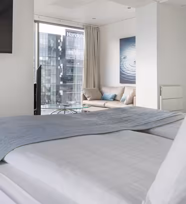 Modern hotel room with a neatly made bed covered in white and gray bedding. A TV is mounted on the wall. Large windows reveal a city view with a building labeled Handels. A couch sits in the corner beside a serene water-themed artwork.