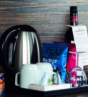 Close-up of a hotel room refreshment tray with a stainless steel electric kettle, tea and coffee packets, two white mugs, and upside-down wine glasses. Theres a bottle of wine with a menu tag, a can of nuts, and a bag of sea salt chips.