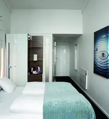 A modern hotel room with a neatly made bed featuring white linens and a light blue throw. A large, abstract art piece hangs on the wall. A flat-screen TV is mounted opposite the bed, and a narrow corridor leads to the bathroom with an open door.