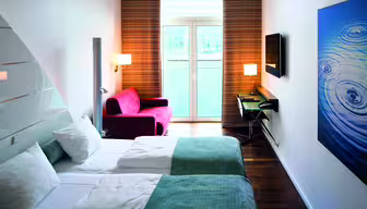 A modern hotel room with two neatly made twin beds, white and blue linens, a red armchair by tall windows, and a wall-mounted TV. A blue artwork with ripple patterns decorates the wall. A small desk and chair are near the window.