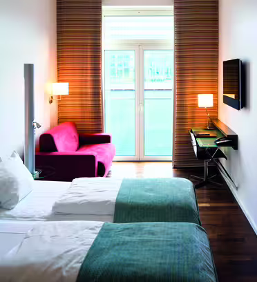 A modern hotel room with two neatly made twin beds, white and blue linens, a red armchair by tall windows, and a wall-mounted TV. A blue artwork with ripple patterns decorates the wall. A small desk and chair are near the window.