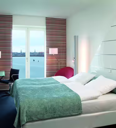 Modern hotel room with a large bed covered in a light blue and white bedspread. A glass desk and chair are near a wall-mounted TV. Windows reveal a scenic waterfront view. The décor includes a red chair and striped curtains.