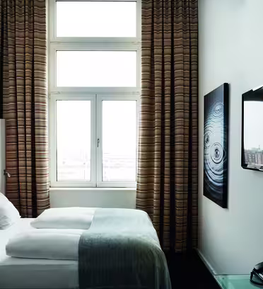 A modern hotel room with a neatly made bed by tall, patterned curtains. A wall-mounted TV is on the right, above a small desk with a phone. A framed abstract artwork hangs next to the window, offering a view of buildings outside.