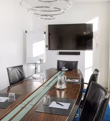 A modern conference room with a long oval table surrounded by black leather chairs. On the table are notepads, pens, and water glasses. A flat-screen TV and flipchart stand on the far wall. Large windows let in natural light.
