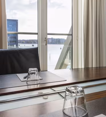 A modern meeting room with a wooden table, empty glass, pen, and notepad, overlooking a balcony with a view of water and distant buildings through glass doors. Cream curtains are slightly open, letting in natural light.