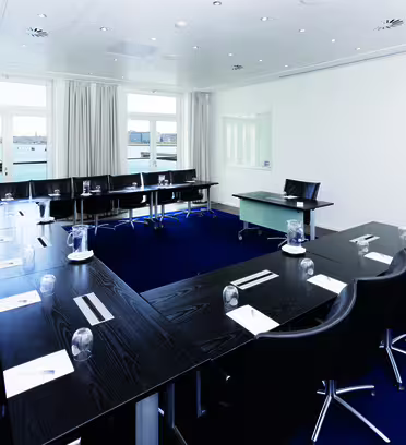 A modern conference room with a long, U-shaped dark wood table surrounded by black chairs. Notepads and water glasses are placed on the table. Large windows offer a view of water and buildings outside. White walls and blue carpet complete the setting.
