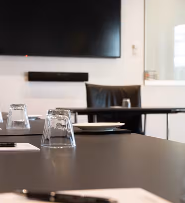 A modern conference room with a large flat-screen TV on the wall. A long table is set with upside-down glasses, pens, and notepads. There is a chair at the end of the table. The room has white walls and a clean, professional look.